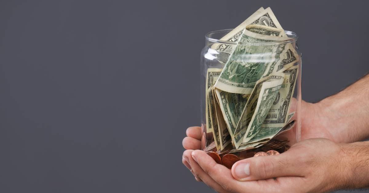 A man is holding a glass donation jar in his outstretched hands. The jar is filled with coins and bills.