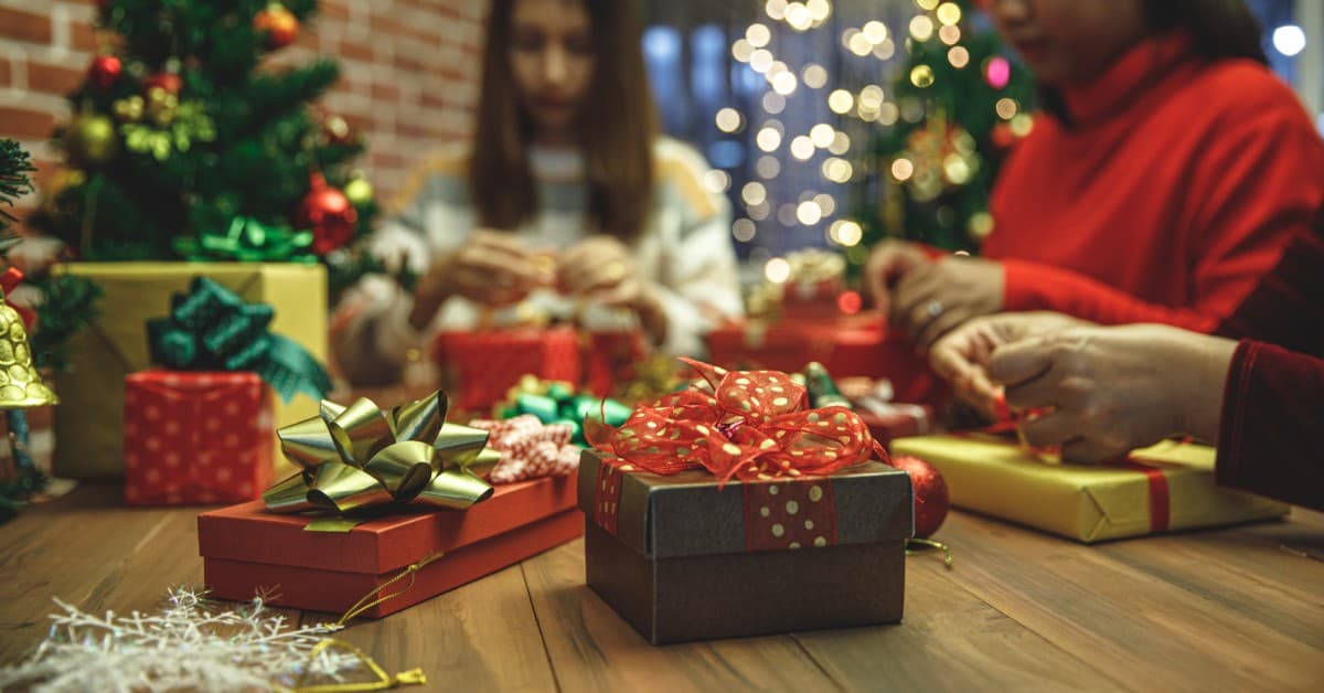 Three people are sitting at a table with a Christmas tree in the background. They're wrapping gifts with bows and ribbons.