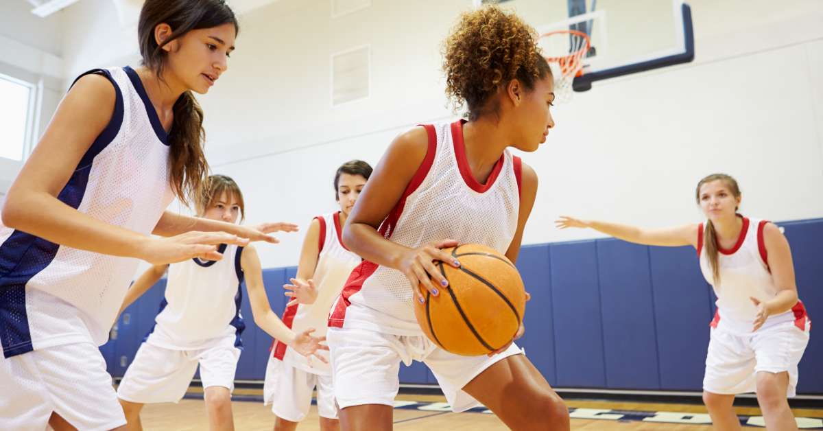 Five young females playing in a basketball game. One individual is holding the ball while the other players surround her.