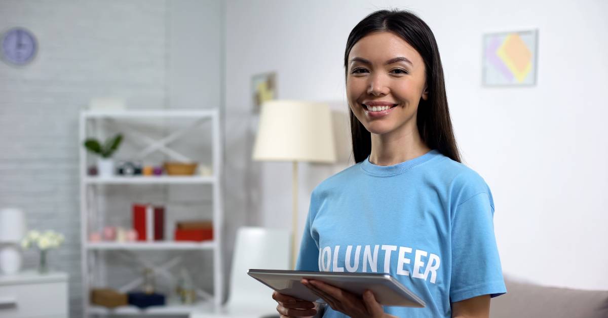 A young woman wearing a light blue T-shirt that says "Volunteer" in white letters. She is smiling and holding a tablet.