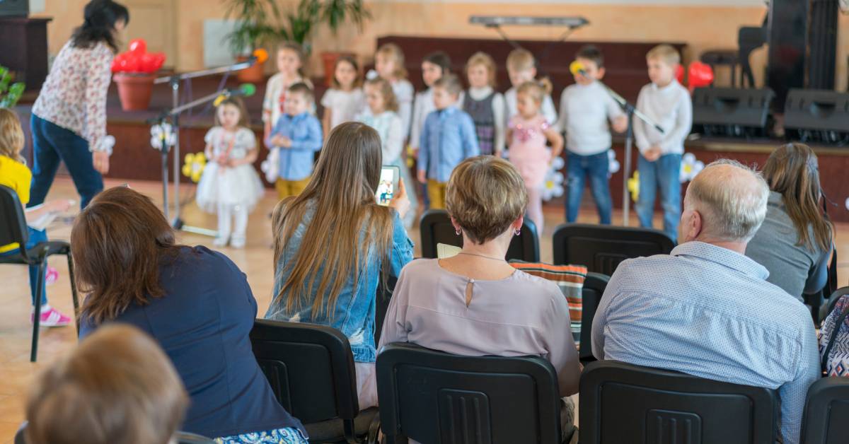 A group of young boys and girls performing in front of an adult audience. One adult is taking a photo with her phone.