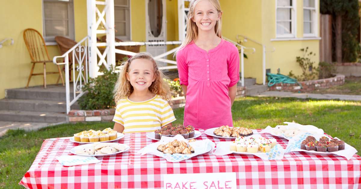 Two young girls standing behind a bake sale table filled with sweet treats. The table is located outside a yellow house.