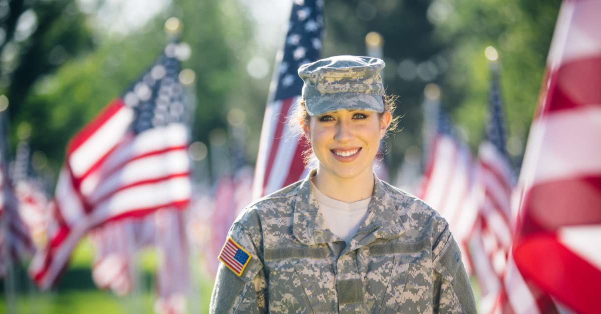 A female soldier dressed in a military uniform standing outside. There are several American flags behind her.