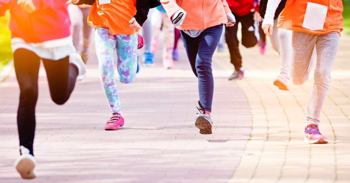 A group of children are running outside on a sunny day. They are wearing race bibs on the front of their shirts.