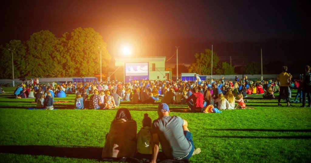 A group of people are watching an outdoor movie at night. They are sitting on blankets on a large field.