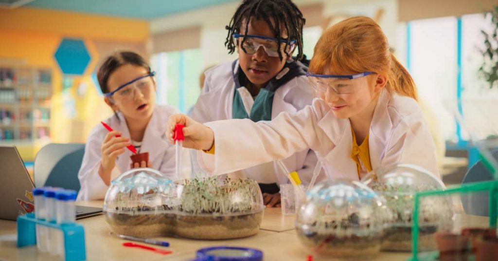 Three young students are performing a science experiment. They are wearing lab coats and protective glasses.