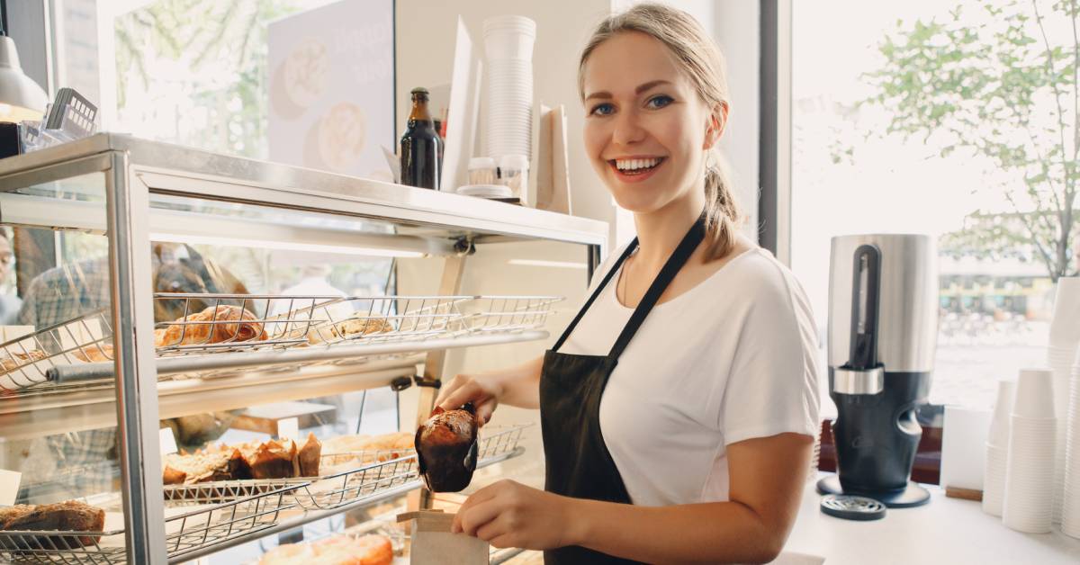 A female employee is wearing a black apron and standing behind a bakery case. She is putting a muffin in a bag.