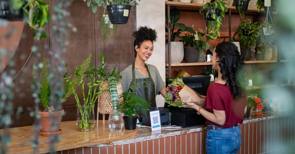 A woman is selling a bouquet of flowers to a customer in a plant shop. She is wearing an apron and smiling at the customer.