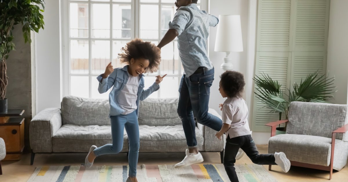 A man, a young girl, and a young boy are smiling, jumping, and dancing in a circle in a living room.