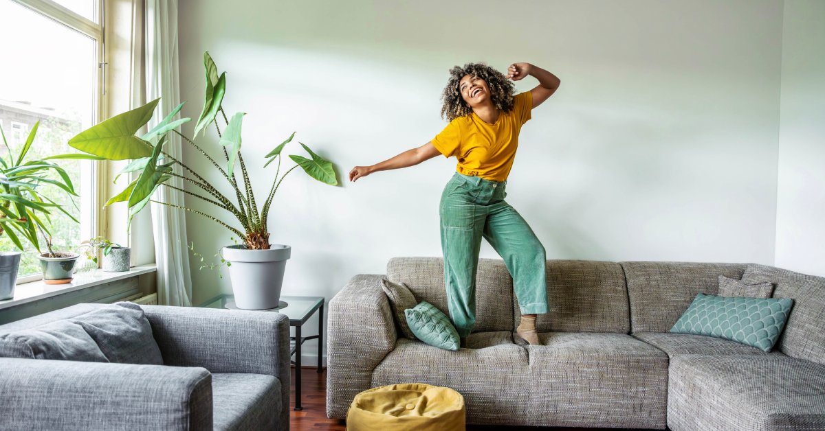 A woman wearing green pants and a gold shirt is standing on a tan couch and smiling. She is dancing by herself on the couch.