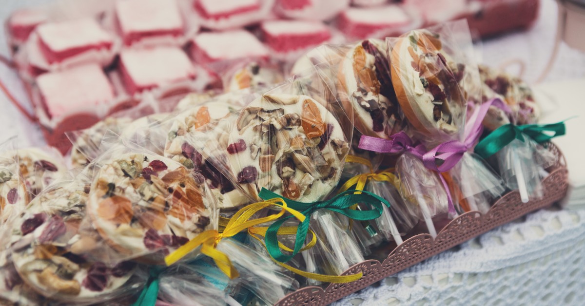 A tray of lollipops are individually packaged and tied with a ribbon. There is another tray of desserts in the background.