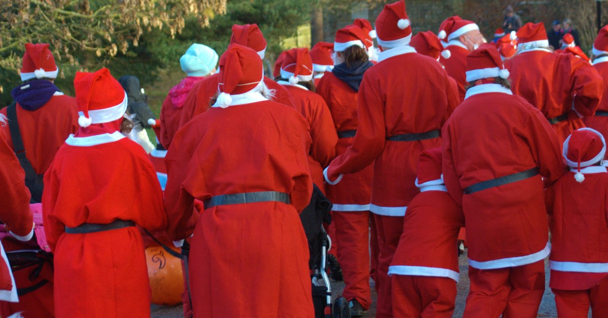 A group of people dressed in red and white Santa costumes that are walking together outside. Their backs are to the camera.