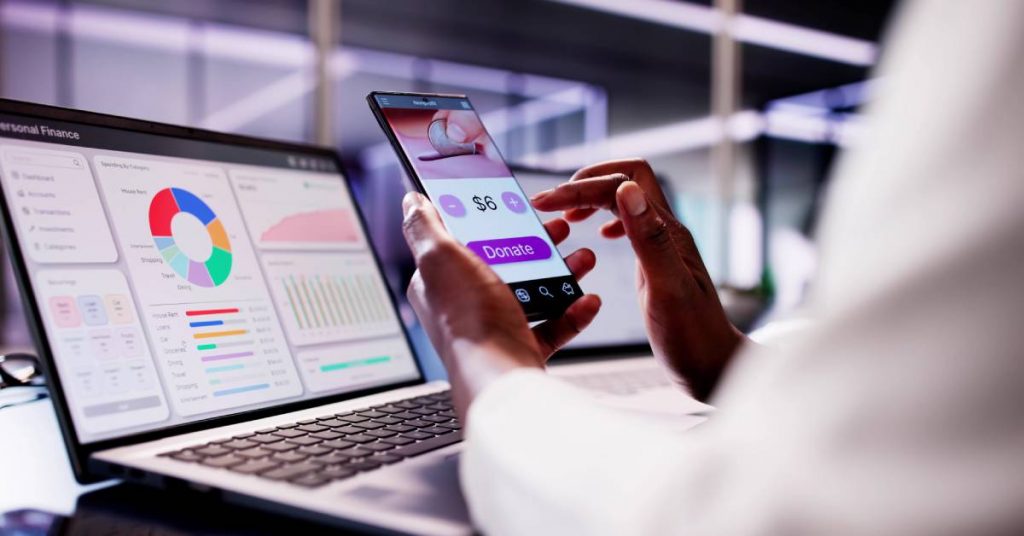 A person holding a smartphone and looking at a donation screen. There is a laptop with graphs and charts behind the phone.