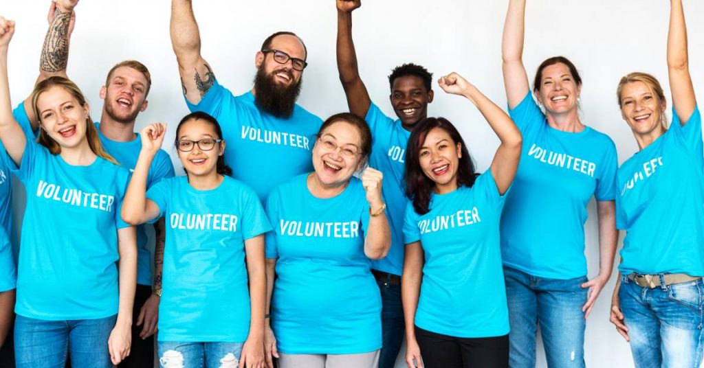 Several people wearing blue t-shirts with "volunteer" written on the chest. They are happily raising their arms in the air.