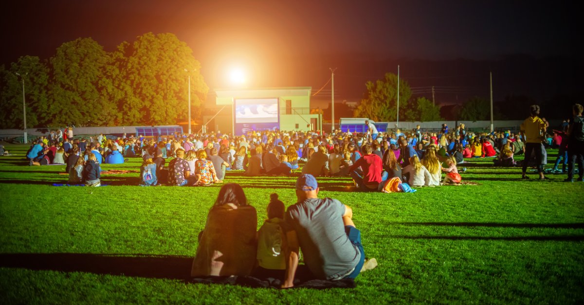 A large group of people are sitting on blankets in a grassy field and watching a movie on a screen at night.