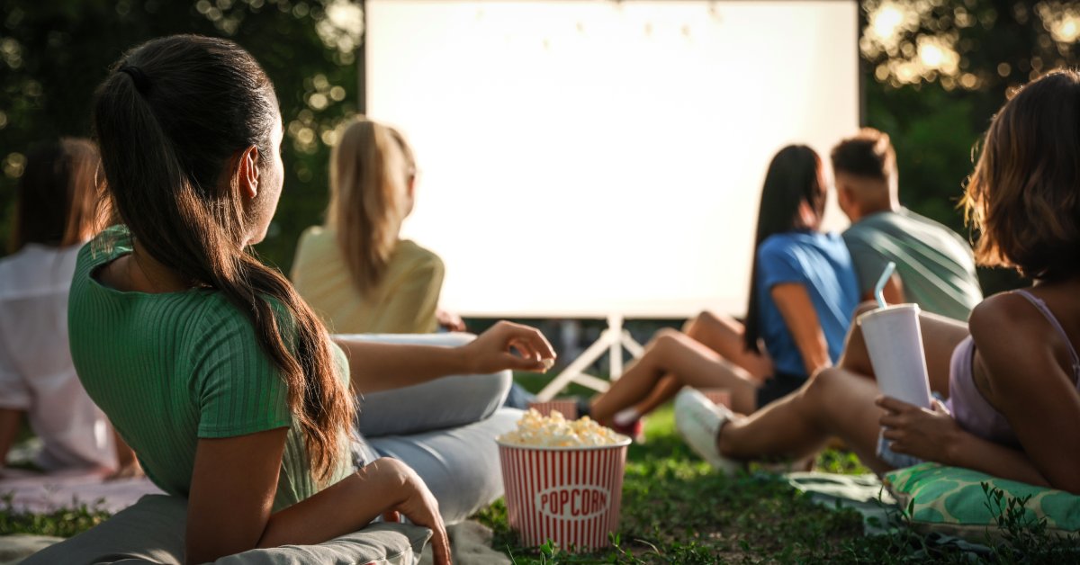 A group of young people are sitting on the ground and watching a movie on an outdoor projector while eating popcorn.