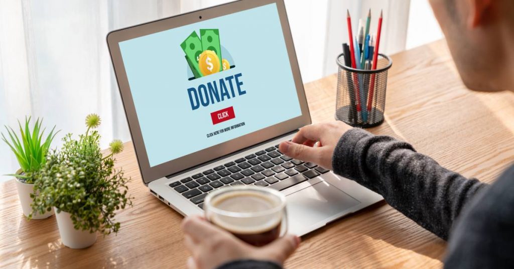 A man is sitting in front of an open laptop and looking at a donation site. He is holding a beverage in one hand.