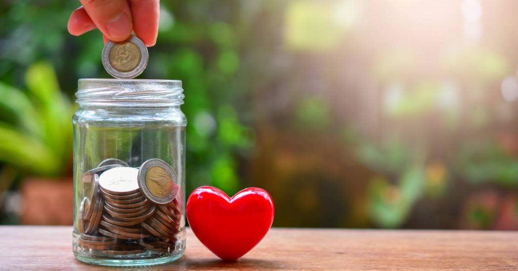 An individual is dropping a coin into a glass jar filled with coins. There is a red heart next to the jar.