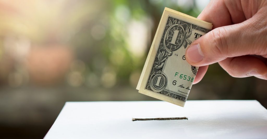 An individual is placing a United States one-dollar bill into a white box with a small opening on the top.