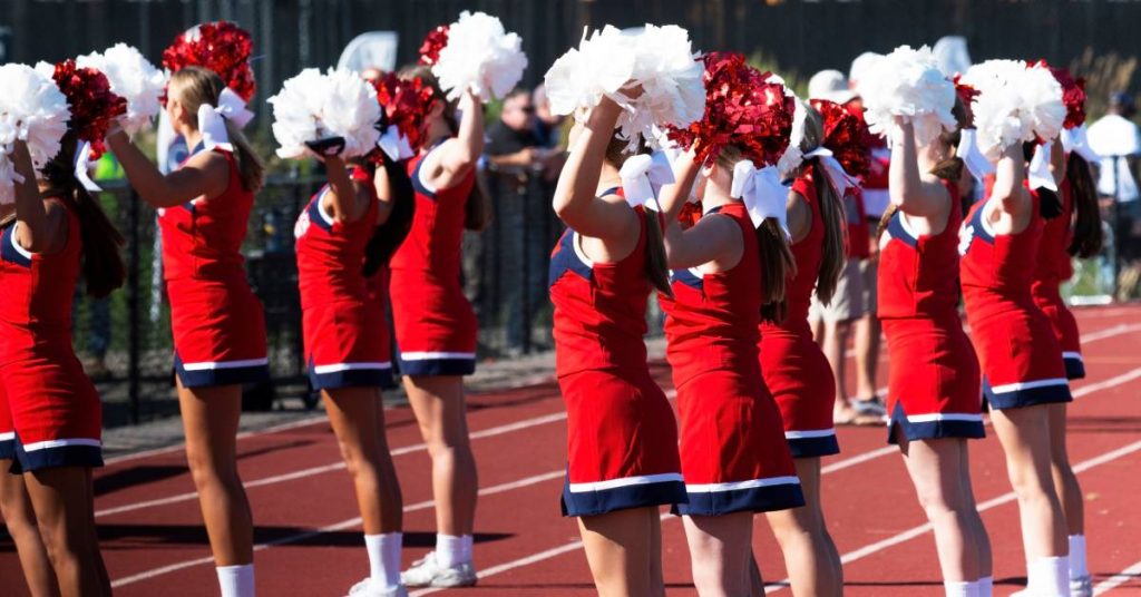 A group of cheerleaders wearing red uniforms are holding their pom poms in the air while performing outside.