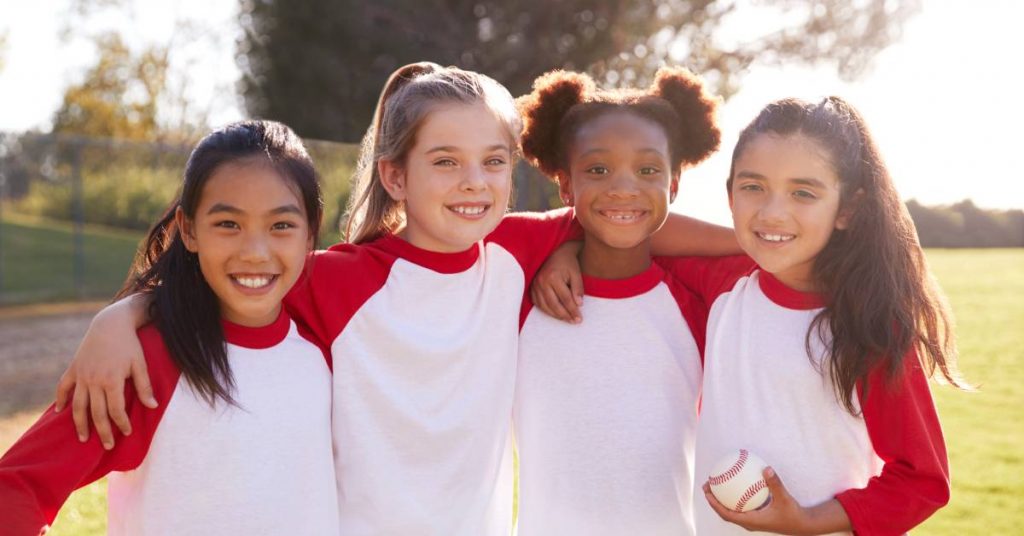  A group of young girls are wearing red and white shirts and embracing one another. One girl is holding a baseball.