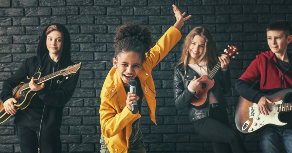 Four young people playing music together. Three people are playing guitar and one is singing into a microphone.