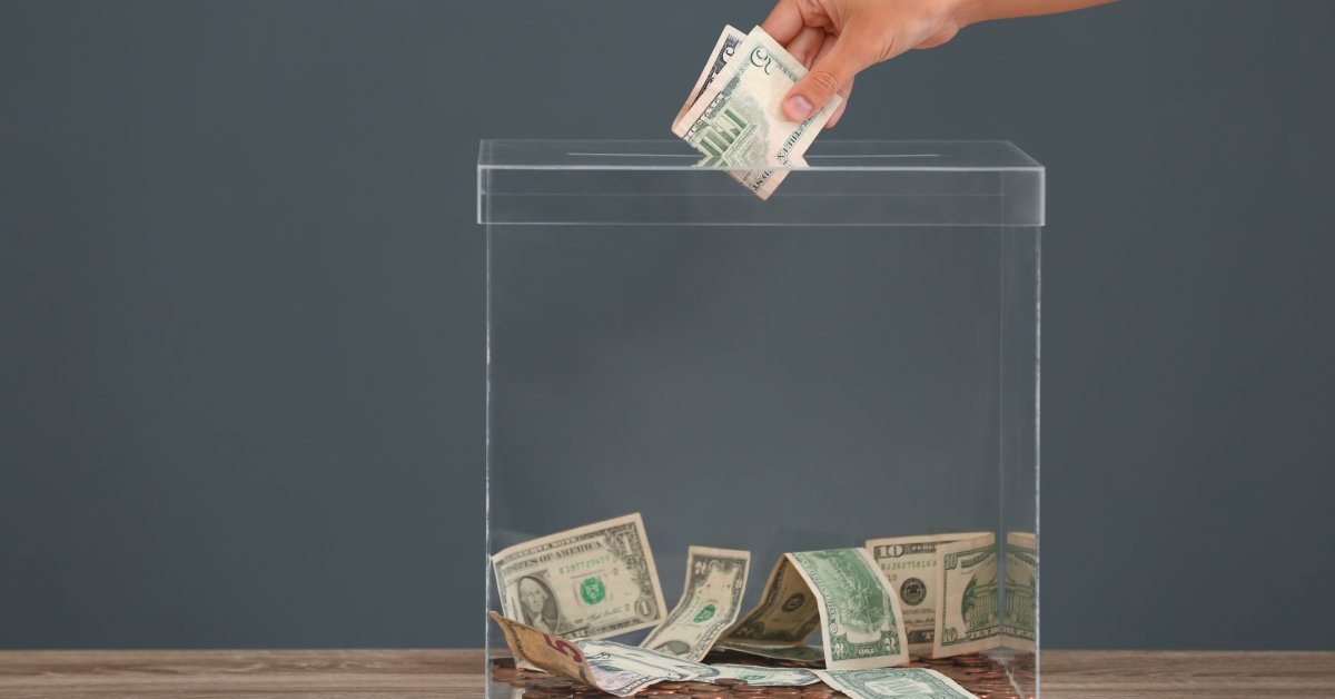 A person putting a five-dollar bill into a clear box filled with bills and coins. The box is sitting on a table.