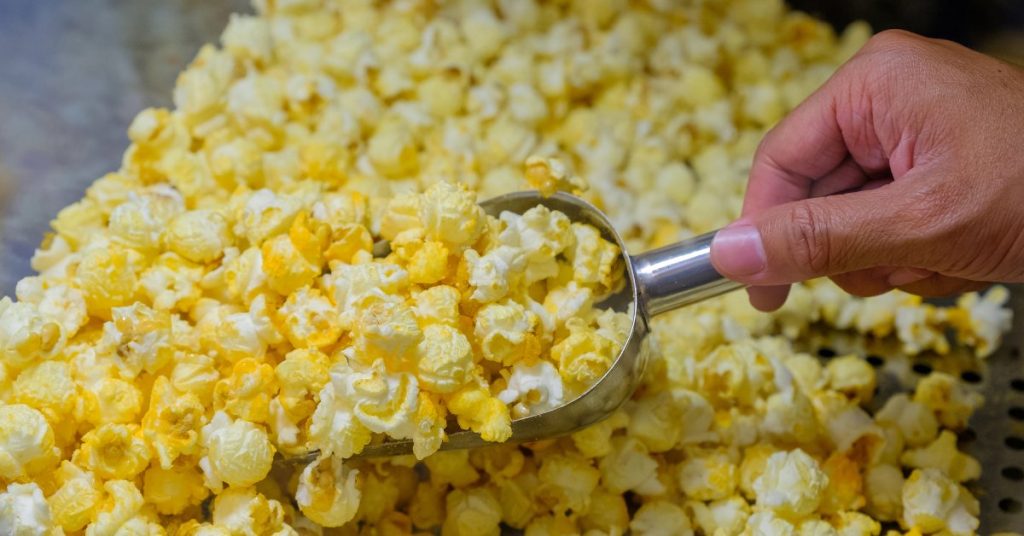 A close-up of a person using a silver scoop to pick up yellow and white pieces of popcorn from a large pile.