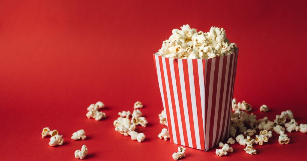 A red and white striped box filled with popcorn on a red background. Some pieces of popcorn have fallen out of the box.