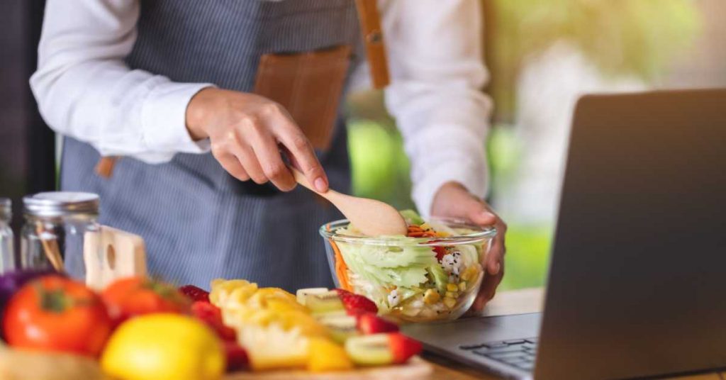 A person wearing a striped apron is making a colorful salad in a glass bowl while looking at a laptop.