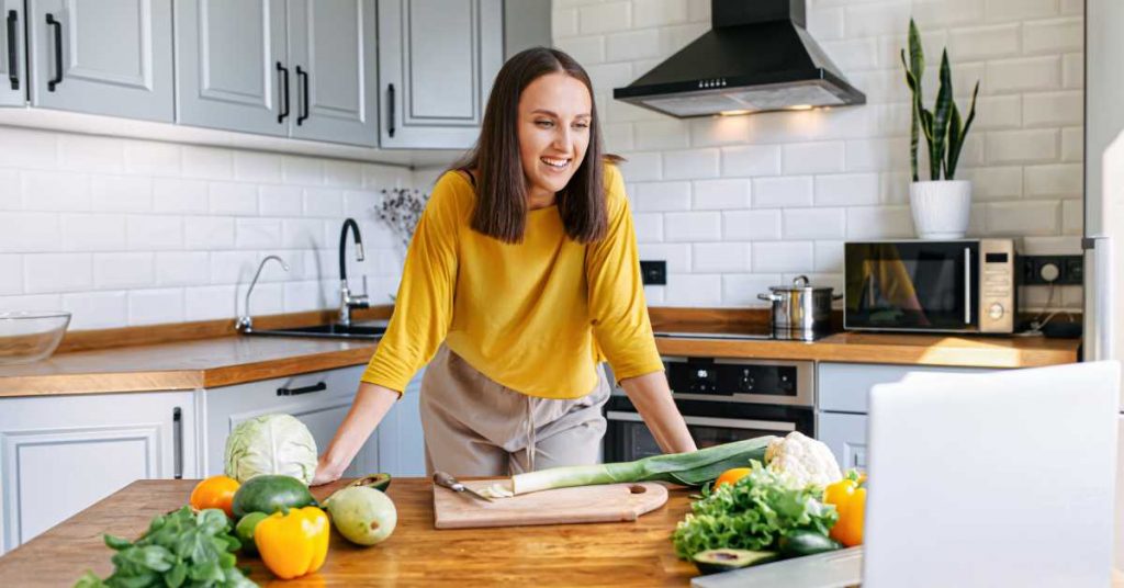 A young woman is wearing a yellow shirt and cutting vegetables in her kitchen while looking at a laptop.